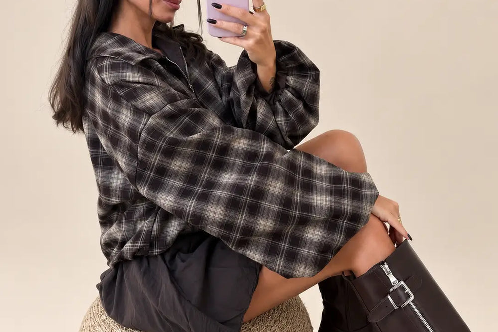 Frau sitzt auf einem geflochtenen Hocker in weiter karierter Hemdjacke in Braun mit Reißverschluss und Ballonärmeln, dazu dunkle Shorts und hohe braune Stiefel | Lisa’s Boutique