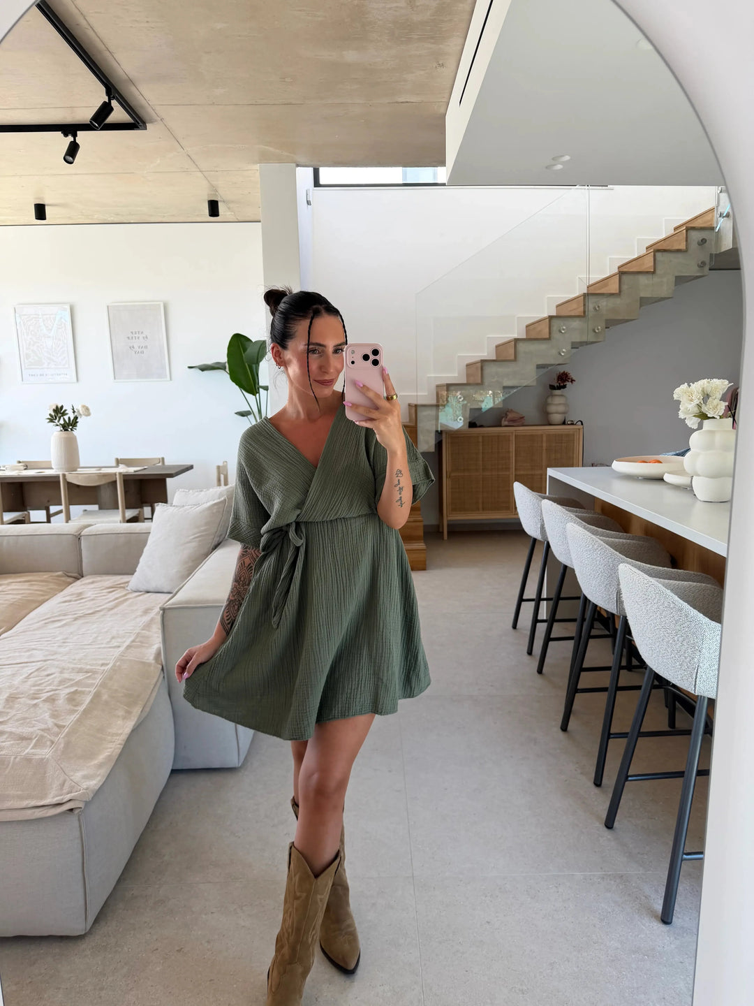 Frau in einem kurzen Musselinkleid in Khaki mit weitem Ausschnitt, Bindeband an der Taille und kurzen weiten Ärmeln, dazu beige Stiefel | Lisa's Boutique