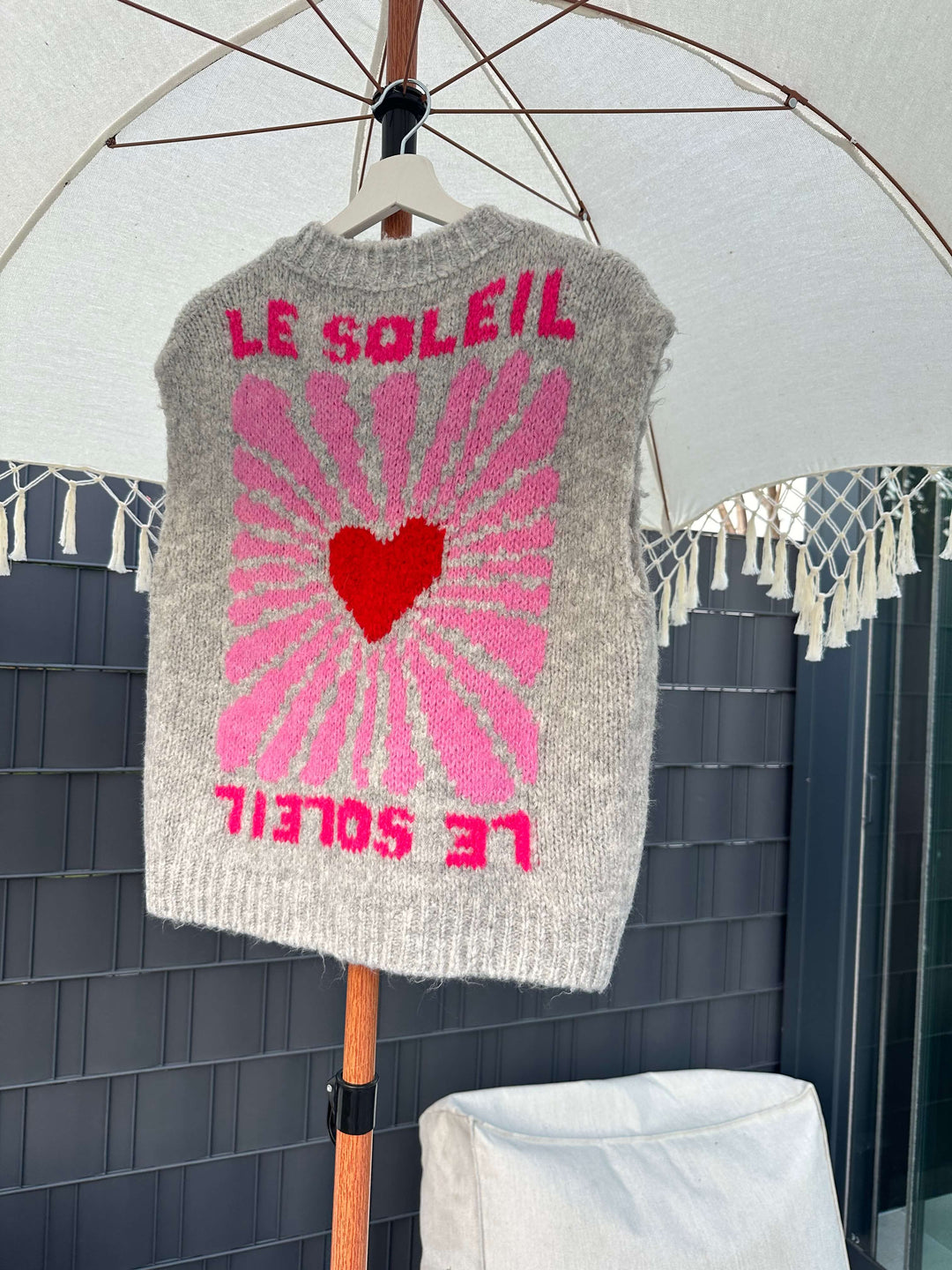 Ärmelloser grauer Strickpullover mit Knopfleiste und pink-rotem „Le Soleil“-Print mit Herzmotiv, hängend auf einem Bügel unter einem weißen Sonnenschirm.