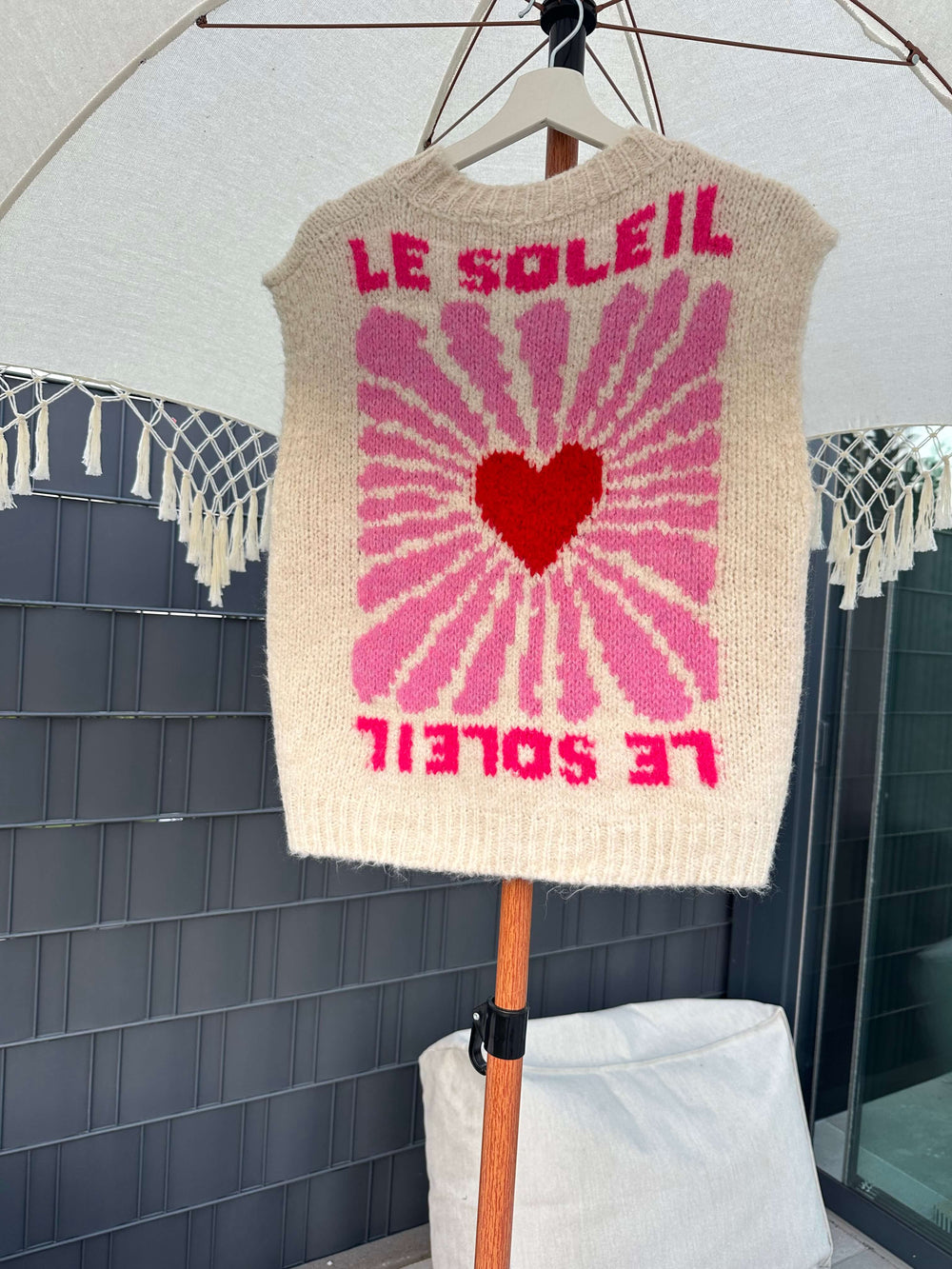 Rückseite eines cremefarbenen ärmellosen Strickpullovers mit großem pinken „Le Soleil“-Print und rotem Herzmotiv in der Mitte, hängend auf einem Bügel unter einem Sonnenschirm.