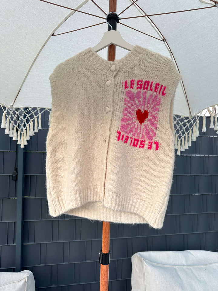 Ärmelloser cremefarbener Strickpullover mit Knopfleiste und pinkem „Le Soleil“-Print mit Herzmotiv, hängend auf einem Bügel unter einem Sonnenschirm.