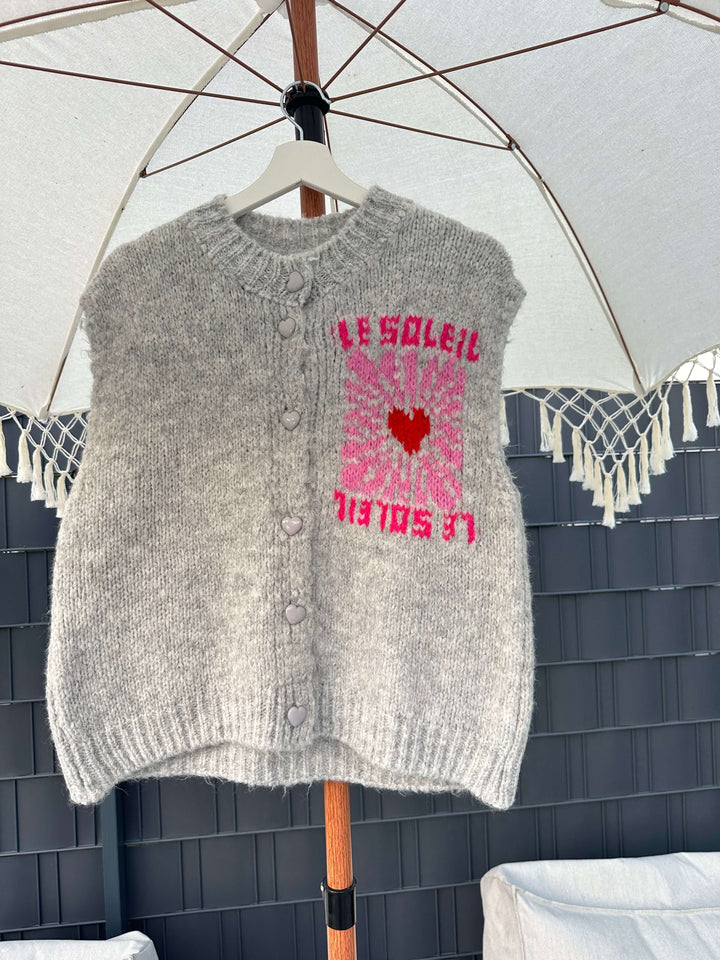 Ärmelloser grauer Strickpullover mit Knopfleiste und pink-rotem „Le Soleil“-Print mit Herzmotiv, hängend auf einem Bügel unter einem weißen Sonnenschirm.