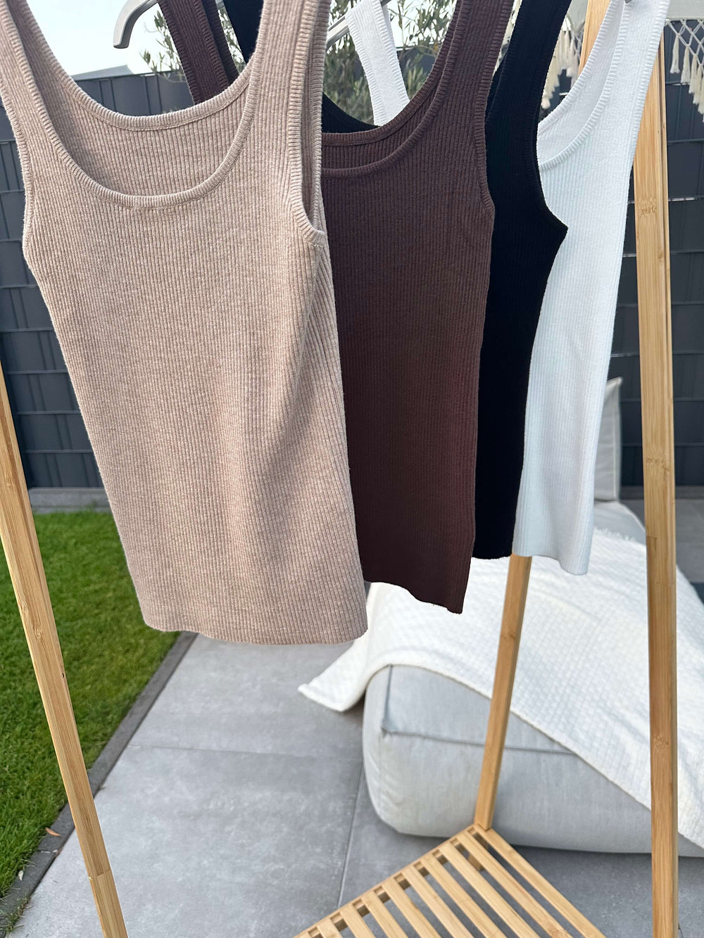 Drei Basic Tanktops in Beige, Schwarz und Weiß hängen auf einem Holzständer im Outdoor-Bereich.