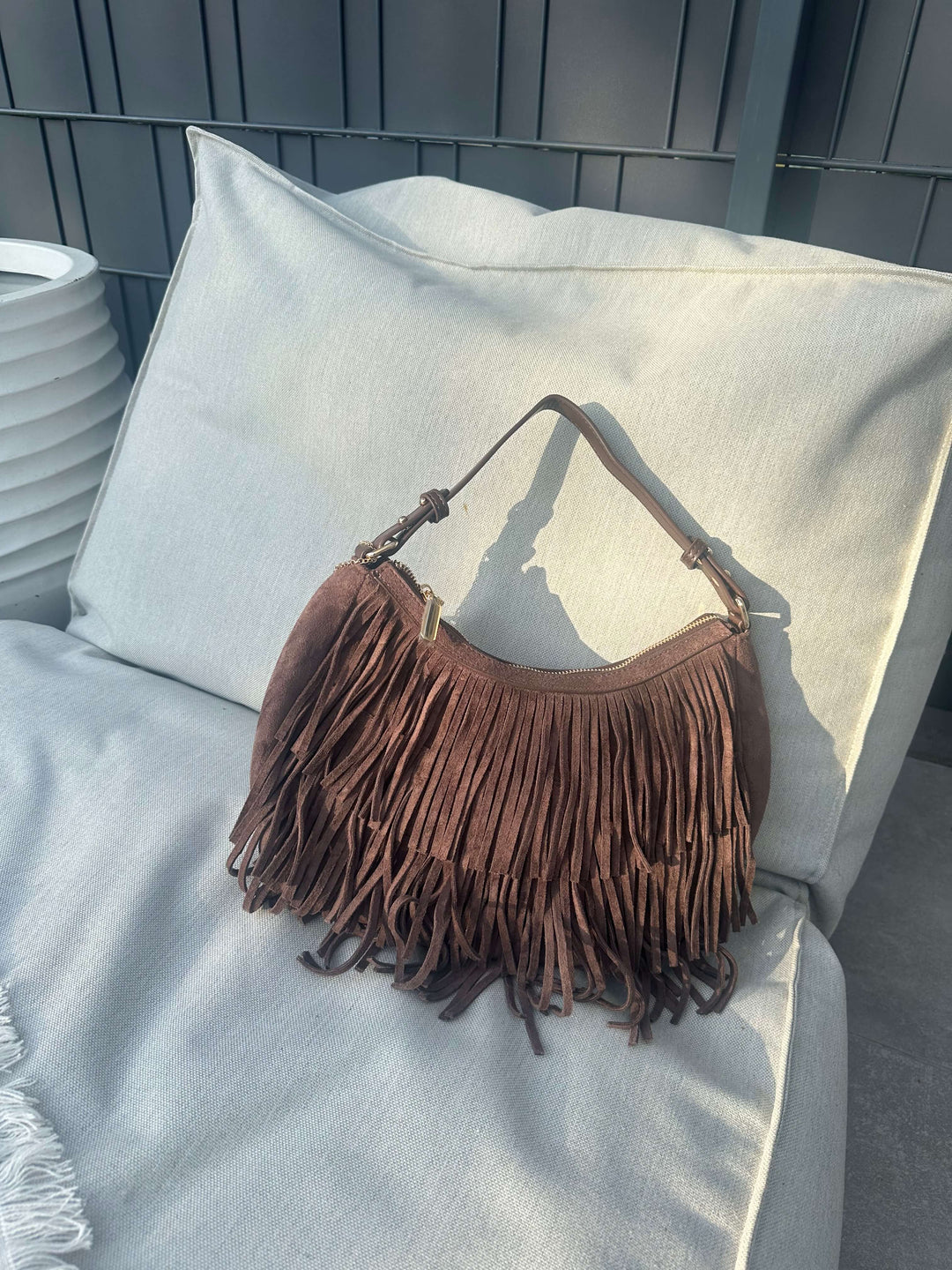  Braune Schultertasche in Wildleder-Optik mit langen Fransen, platziert auf einem hellen Outdoor-Sofa vor einer dunkelgrauen Wand.
