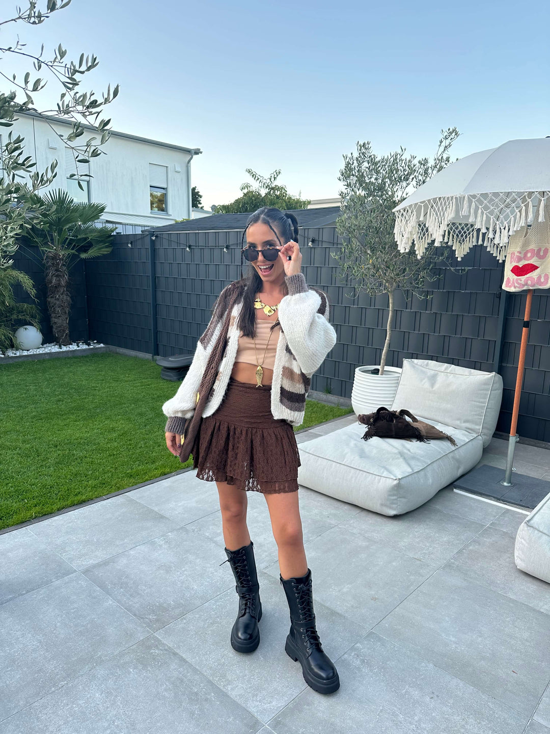 Frau trägt einen braunen, gerafften Minirock mit passendem Bandeau-Top, darüber eine cremefarbene Strickjacke mit braunen Streifen, kombiniert mit schwarzen Stiefeln, goldenen Statement-Ketten und Sonnenbrille im Outdoor-Bereich.