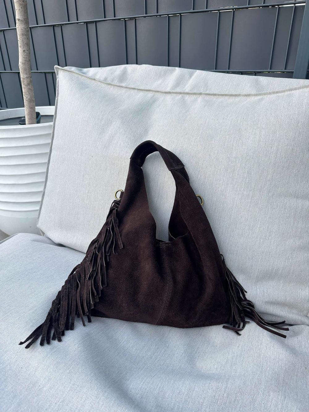 Dunkelbraune Fransen-Schultertasche in Wildlederoptik, liegend auf einem hellen Outdoor-Sofa.