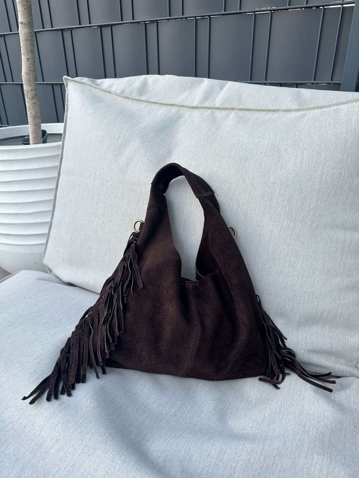 Dunkelbraune Fransen-Schultertasche in Wildlederoptik, liegend auf einem hellen Outdoor-Sofa.