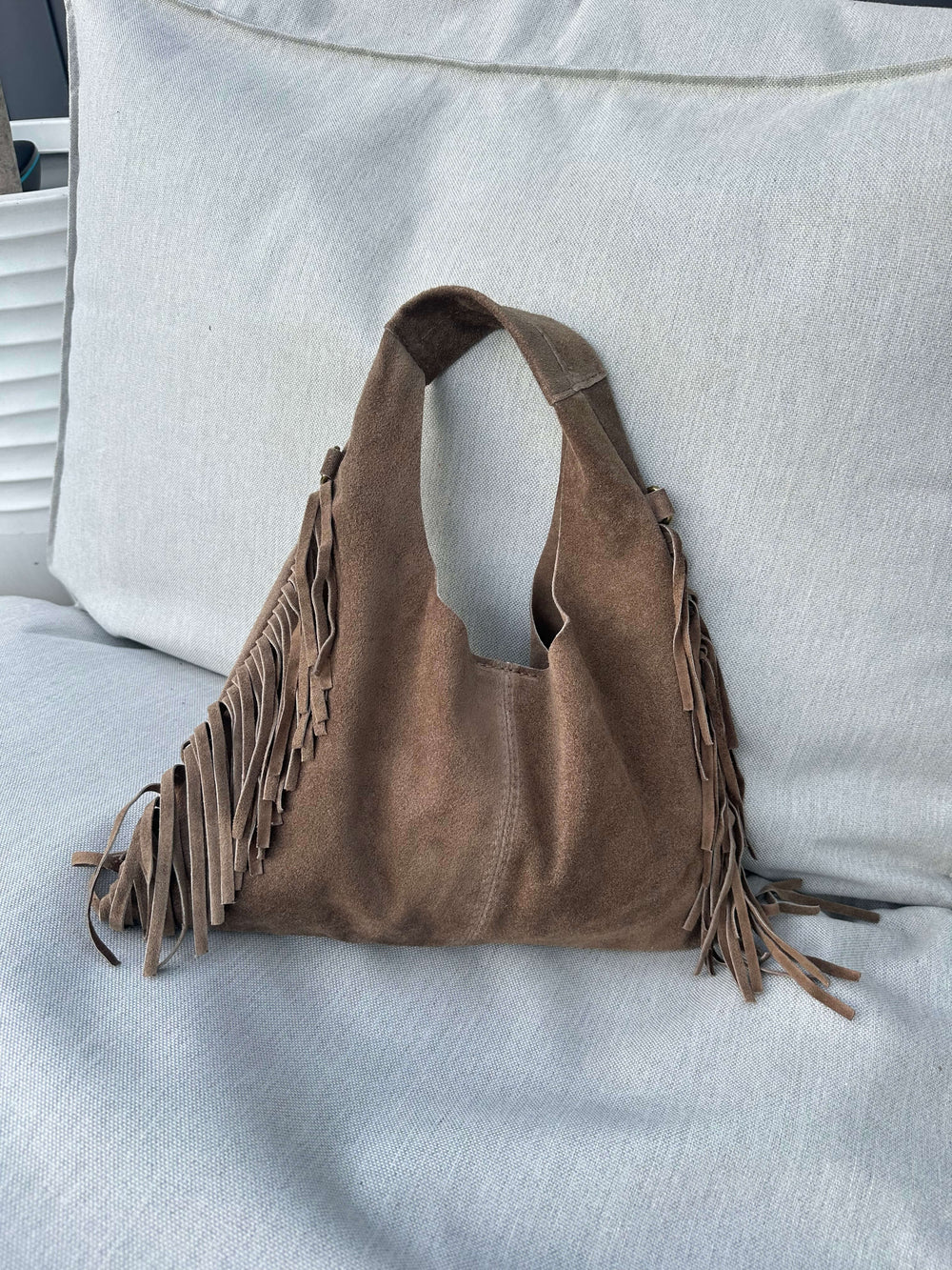 Hellbraune Fransen-Schultertasche in Wildlederoptik, liegend auf einem hellen Outdoor-Sofa.