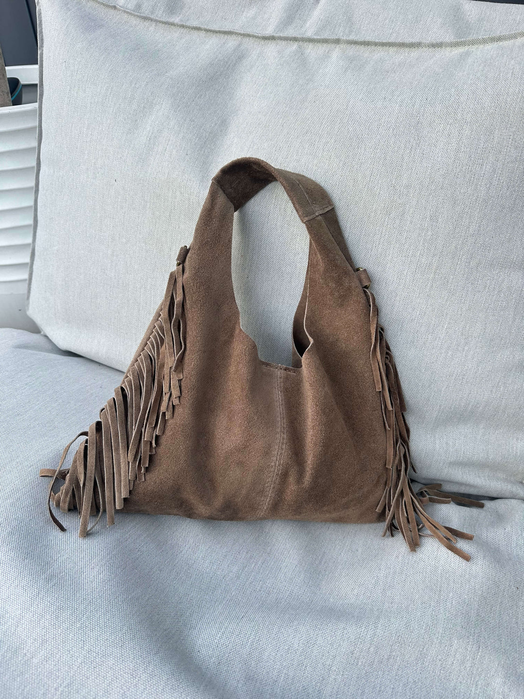Hellbraune Fransen-Schultertasche in Wildlederoptik, liegend auf einem hellen Outdoor-Sofa.