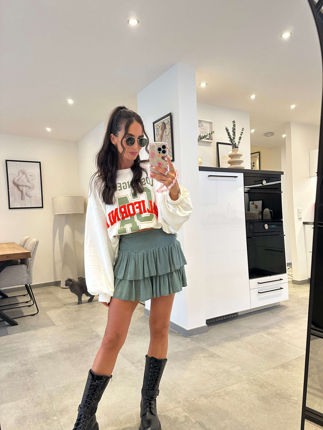 Model trägt weißen Oversize-Pullover mit rotem „California Legendary“-Print und kombiniert ihn mit einem grünen Faltenrock und schwarzen Boots.