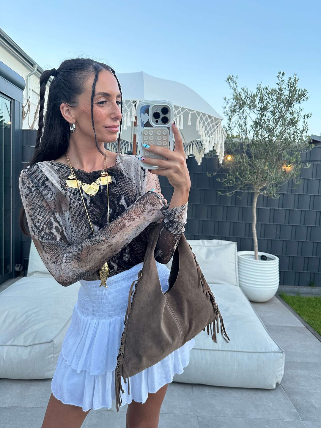 Frau trägt figurbetontes Langarmshirt mit Schlangenmuster in Metallic-Optik, kombiniert mit brauner Fransen-Schultertasche aus Wildlederoptik und weißem Faltenminirock.