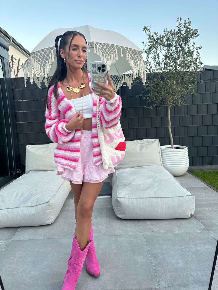 Komplettansicht eines pink-weiß gestreiften Strick-Sets mit passender Strickjacke, Minirock, weißem Crop-Top, pinken Cowboy-Stiefeln und großer Netz-Shopper-Tasche mit rotem Kussmund-Print.