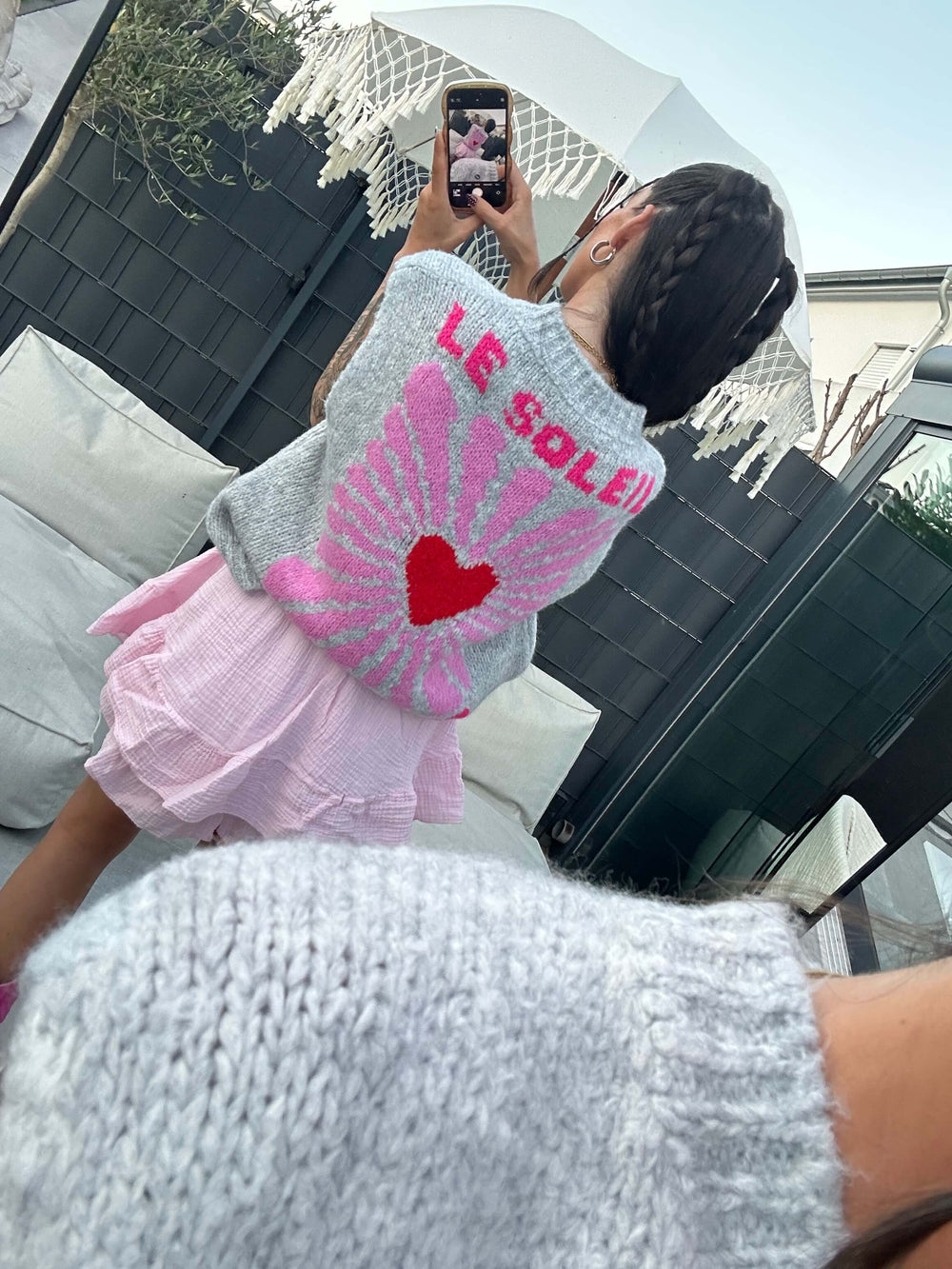 Rückansicht einer Frau in einem grauen Strickpullover mit pinken Flügeln, rotem Herz und der Aufschrift „Le Soleil“, kombiniert mit einem weißen, gewellten Hosenrock.