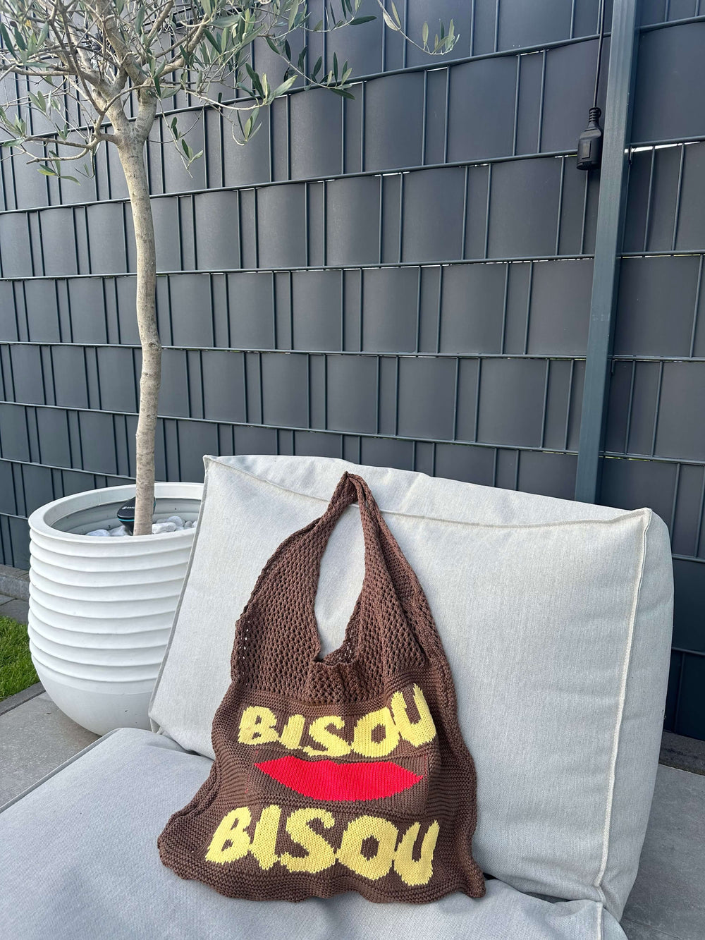 Braune Stricktasche mit gelbem Schriftzug „Bisou“ und rotem Kussmund, liegend auf einem hellen Outdoor-Sofa vor einem Olivenbaum in weißem Blumentopf.