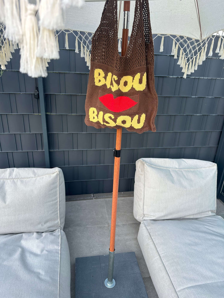 Braune Stricktasche mit gelbem Schriftzug „Bisou“ und rotem Kussmund, hängend an einem Sonnenschirm zwischen zwei hellen Outdoor-Sofas.