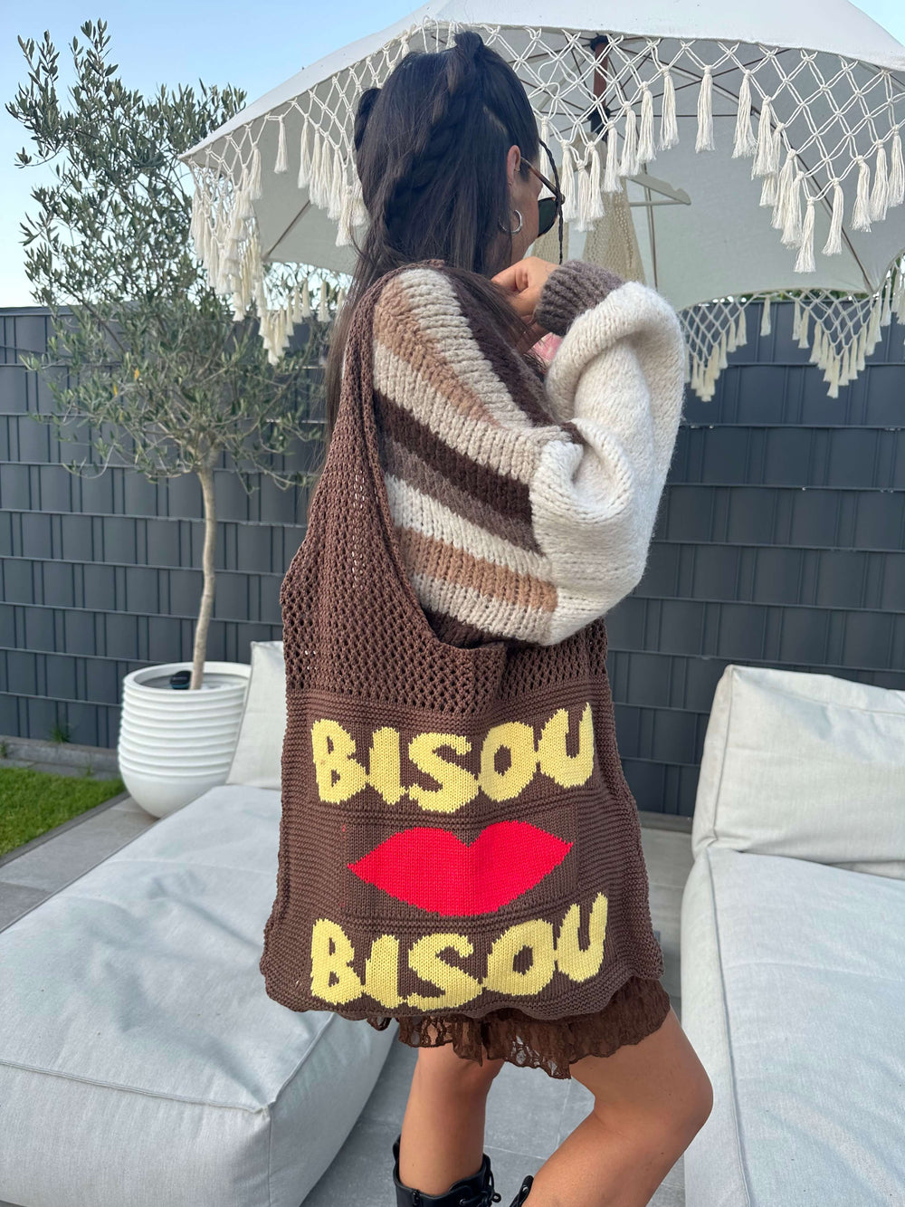 Frau trägt braune Stricktasche mit gelbem Schriftzug „Bisou“ und rotem Kussmund über der Schulter, kombiniert mit braun-beigefarbenem Streifenpullover.