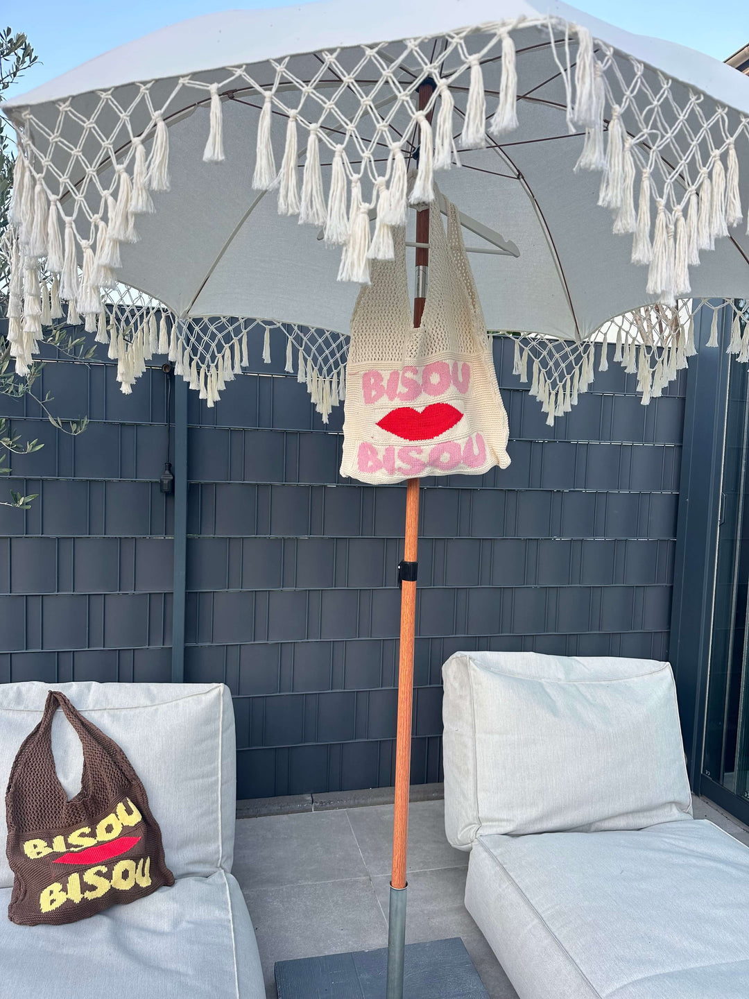 Zwei gestrickte Taschen mit dem Schriftzug „Bisou“ – eine in Creme mit rotem Herz und pinker Schrift, die andere in Braun mit gelber Schrift, dekorativ platziert unter einem weißen Sonnenschirm auf Outdoor-Sofas.
