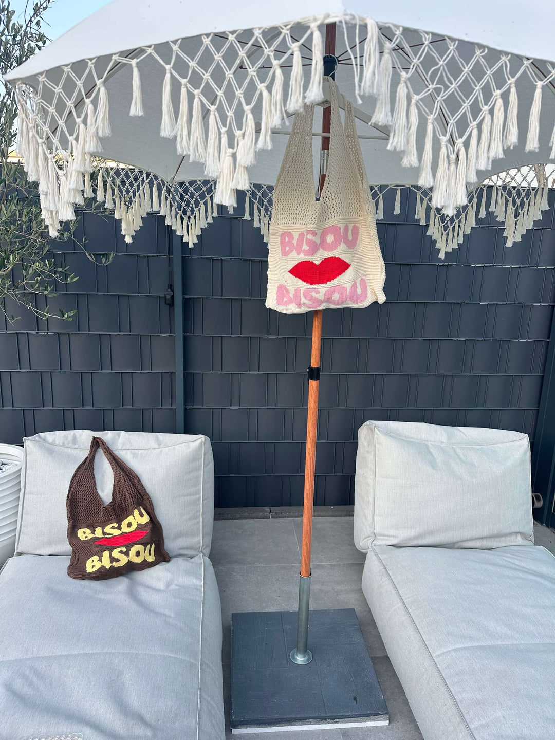 Zwei Stricktaschen mit dem Schriftzug „Bisou“ – eine cremefarbene mit rotem Kussmund und pinker Schrift, hängend am Sonnenschirm, und eine braune mit gelber Schrift, liegend auf einem hellen Outdoor-Sofa.