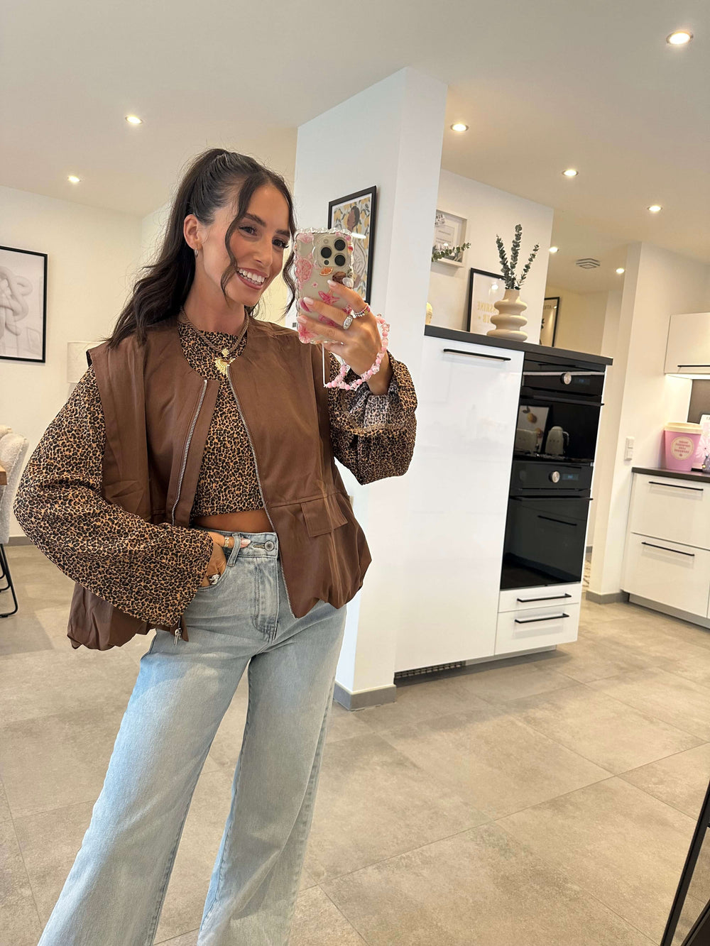 Modell trägt eine braune Weste aus Kunstleder über einer geblümten, langärmeligen Bluse mit Leopardenmuster – kombiniert mit hellblauer High-Waist-Jeans, aufgenommen im modernen Indoor-Ambiente.