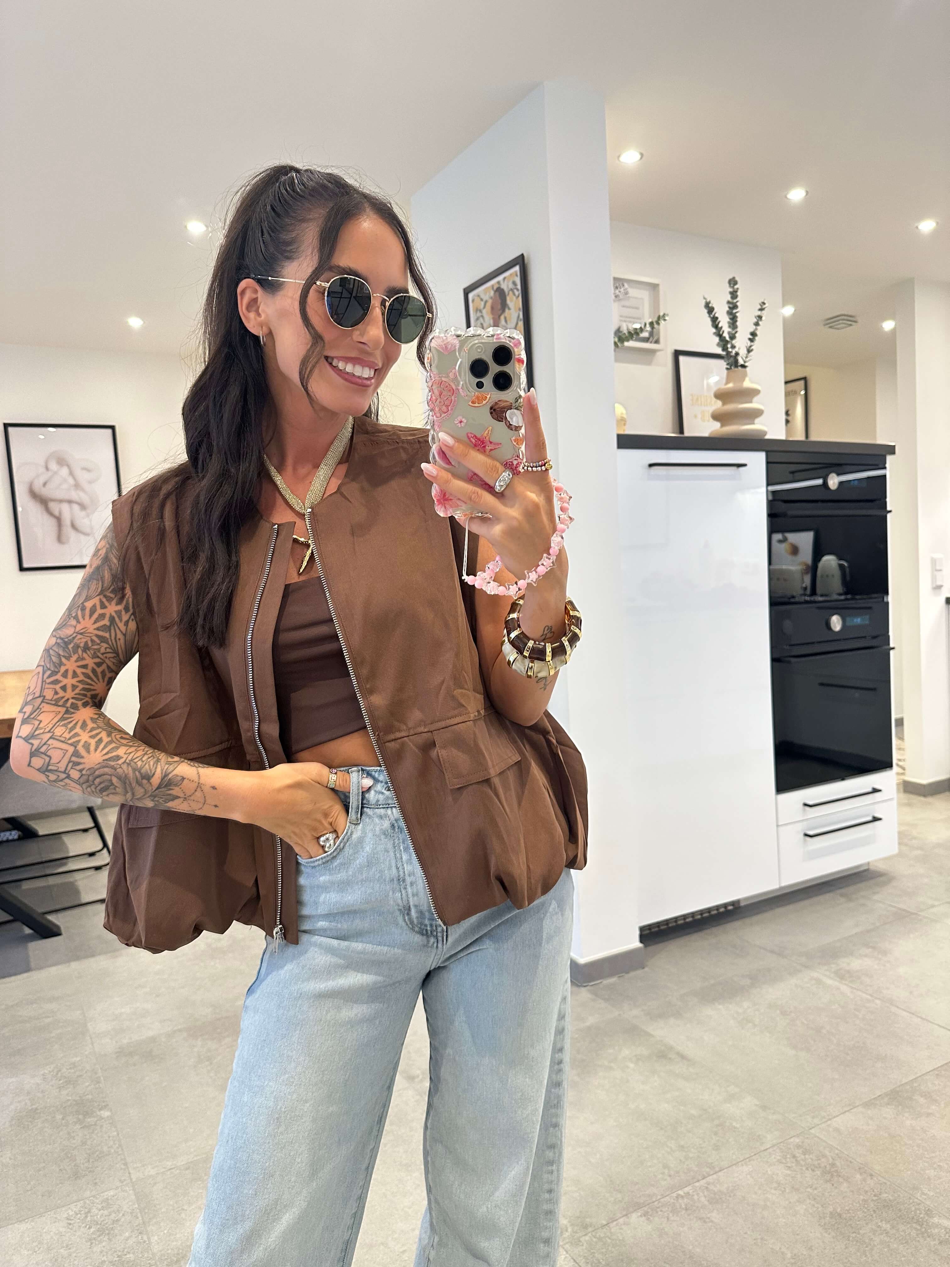 Model trägt eine ärmellose braune Weste über einem schlichten braunen Crop-Top und kombiniert den Look mit einer hellblauen High-Waist-Jeans und Sonnenbrille – moderner, lässiger Style für den Alltag.
