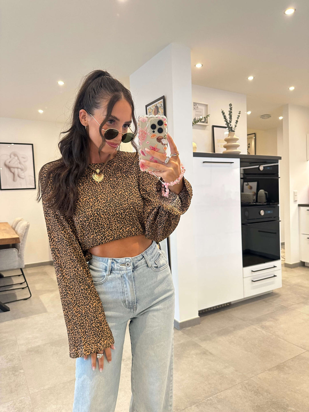 Model trägt ein bauchfreies Leo-Crop-Shirt mit langen Ärmeln – kombiniert mit einer hellblauen High-Waist-Jeans und Sonnenbrille für einen stylischen Look im Alltag.