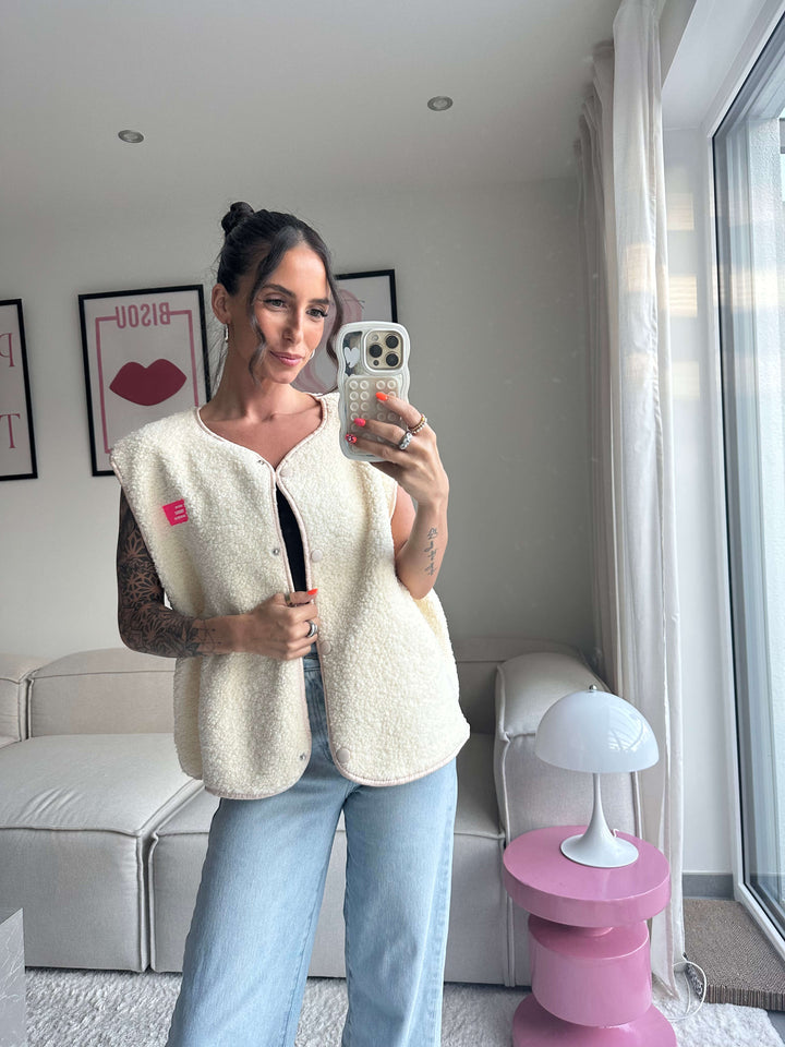 Lisa trägt eine ärmellose cremefarbene Weste aus weichem Flauschstoff mit Knopfverschluss, kombiniert mit schwarzem Oberteil und hellblauer Jeans – Lisas Boutique