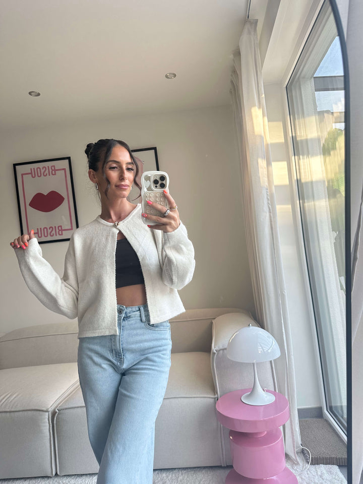 Lisa trägt einen kurzen, cremefarbenen Strickbolero mit Hakenverschluss, kombiniert mit schwarzem Crop-Top und hellblauer Jeans – Lisas Boutique