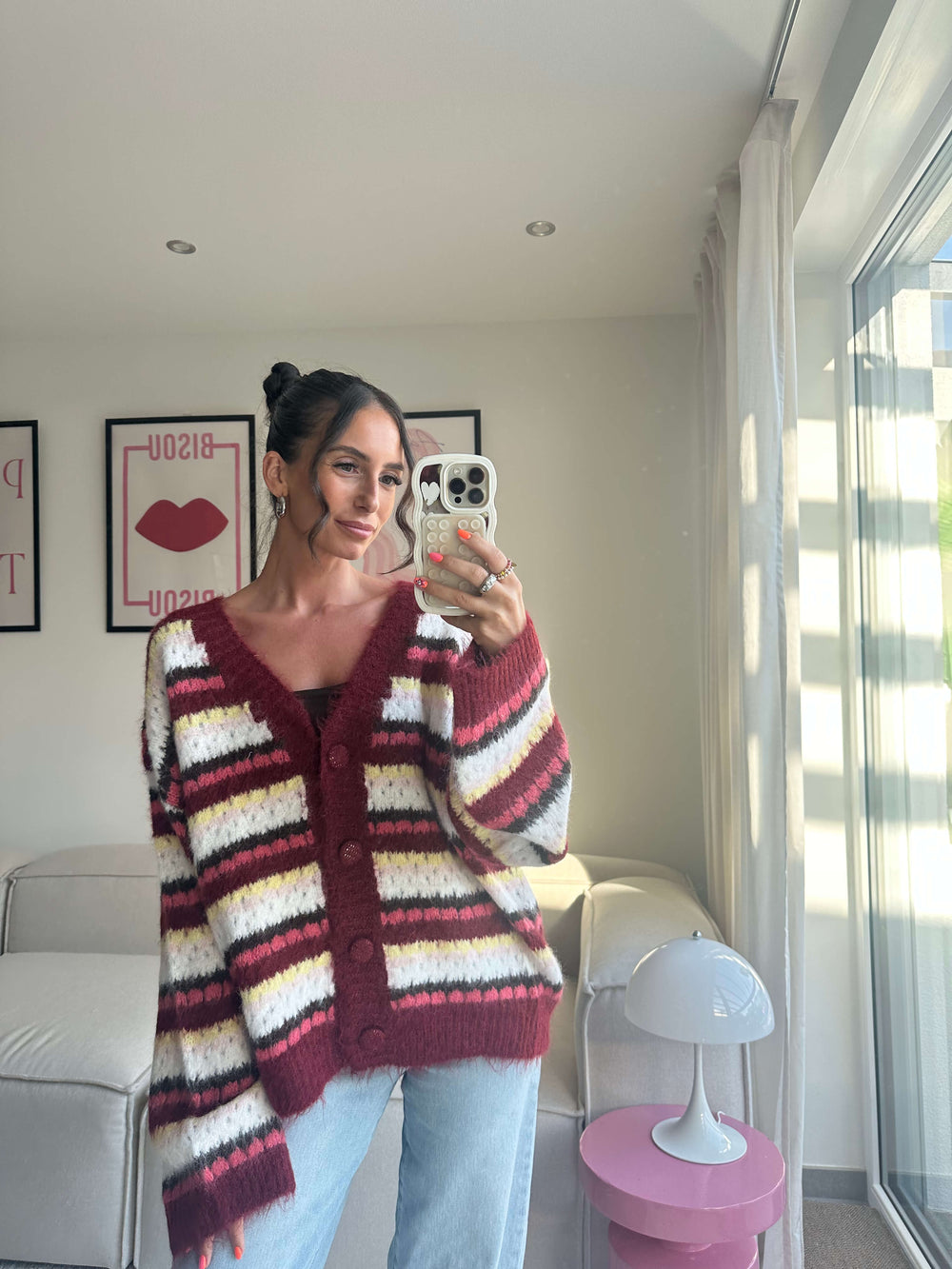 Lisa trägt einen kuscheligen Strickcardigan in Bordeaux mit breiten Streifen in Weiß, Pink, Gelb und Schwarz, kombiniert mit hellblauer Jeans – Lisas Boutique