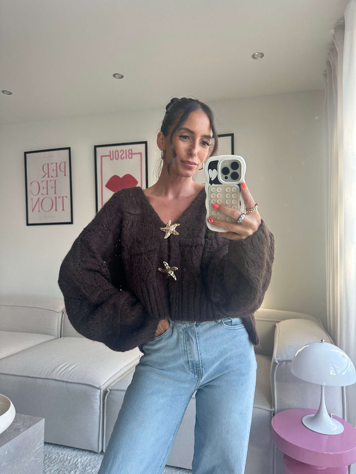 Lisa trägt einen braunen Strickcardigan mit voluminösen Ärmeln und goldenen Broschen, kombiniert mit einer hellblauen Jeans 
