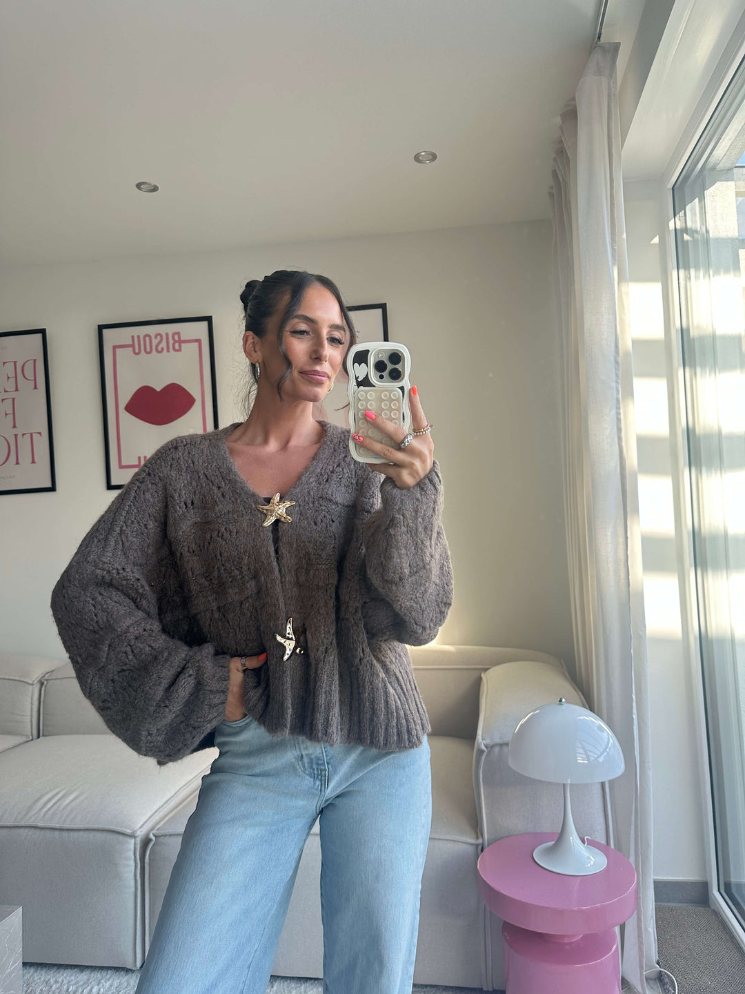 Lisa präsentiert einen braunen Strickcardigan mit weiten Ärmeln und goldfarbenen Broschen, kombiniert mit einer hellblauen Jeans – Boutique