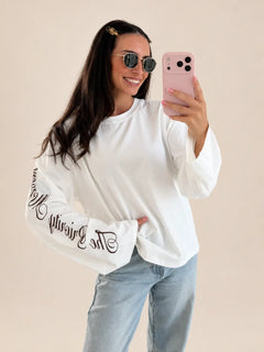 Camiseta manga larga The Priority Moment – Blanca, vista frontal, con texto en la manga, con vaqueros | Lisa’s Boutique