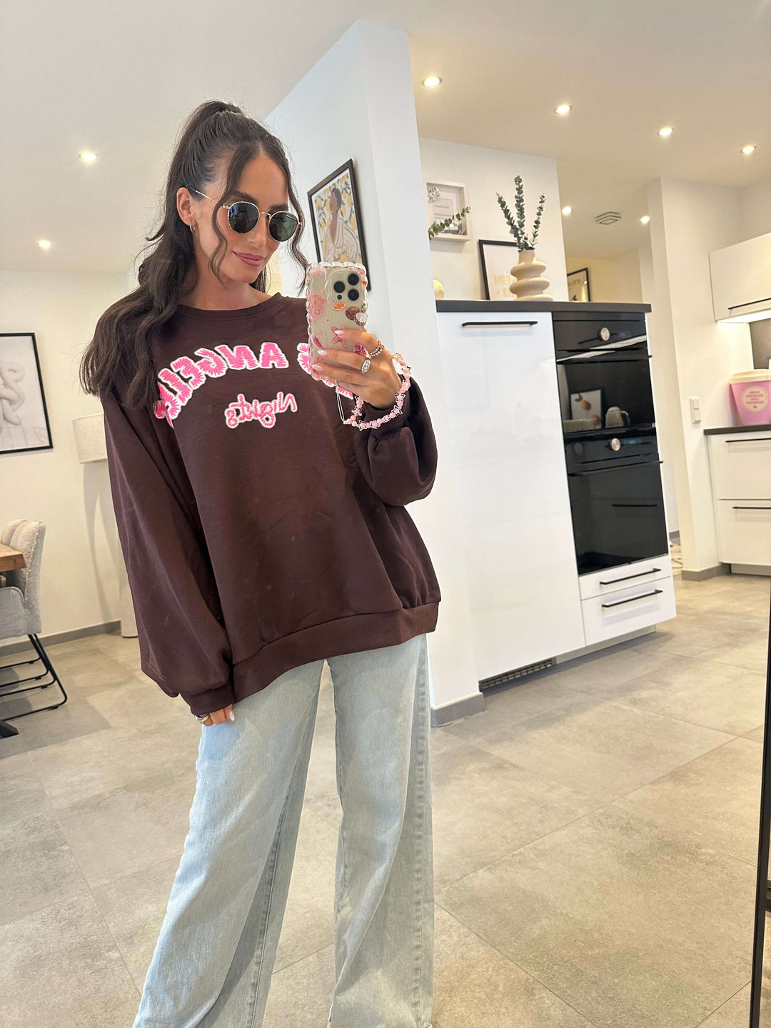 Model trägt einen oversized dunkelbraunen Pullover mit auffälligem „Los Angeles“-Schriftzug in Rot und Weiß, kombiniert mit heller Jeans.