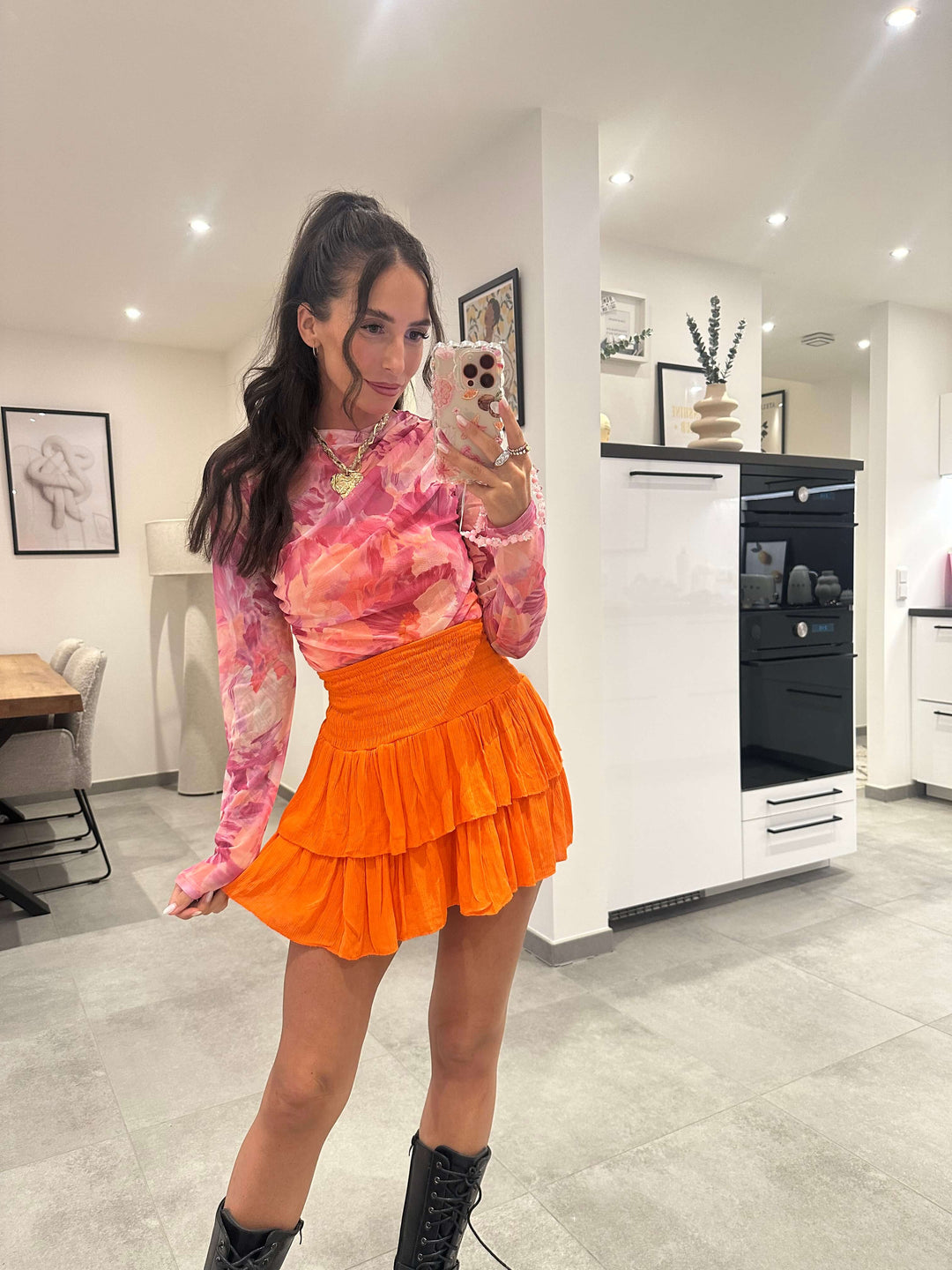 Model trägt ein eng anliegendes, transparentes Mesh-Longsleeve mit pink-orangem Muster und Stehkragen – kombiniert mit einem plissierten Minirock in Orange und schwarzen Boots für einen auffälligen Statement-Look.