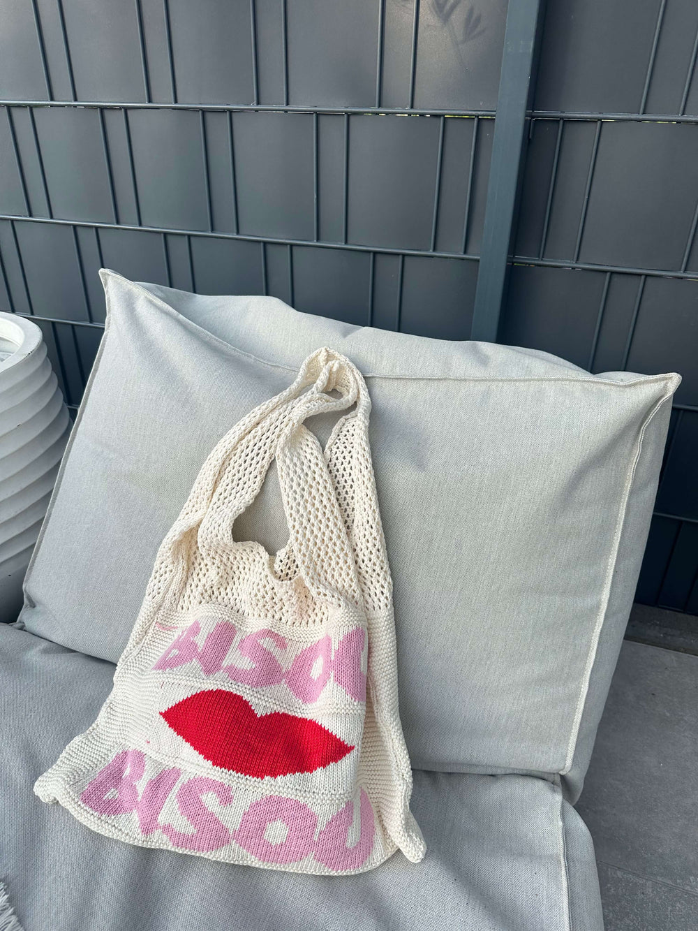 Beigefarbene Netztasche mit rotem Kussmund und pinkem „Bisous“-Schriftzug, platziert auf einem hellen Outdoor-Sofa.