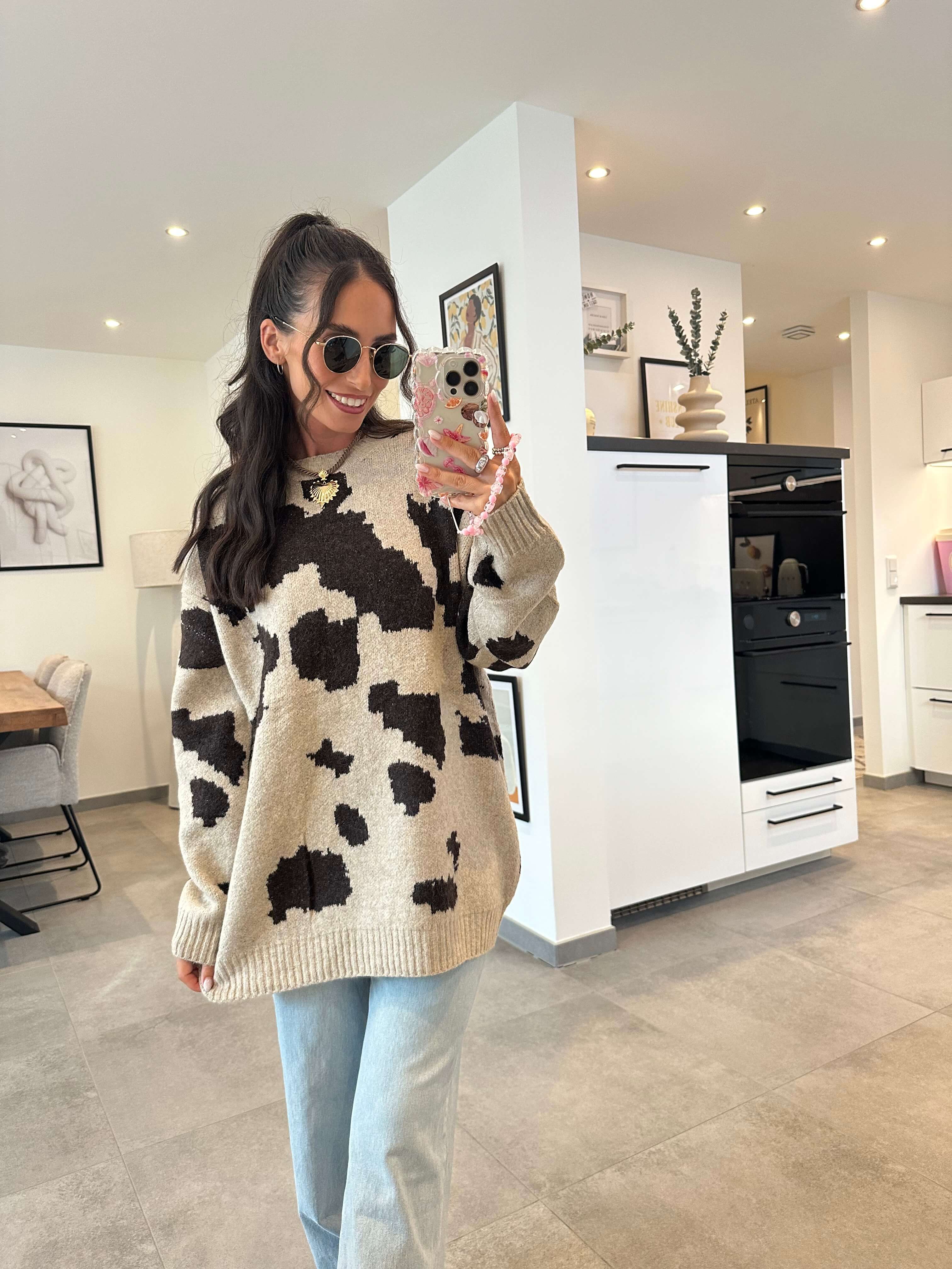 Model trägt einen beigen Oversize-Pullover mit braun-schwarzem Kuhflecken-Muster und kombiniert ihn mit einer hellblauen High-Waist-Jeans – gemütlicher, auffälliger Look für den Alltag.