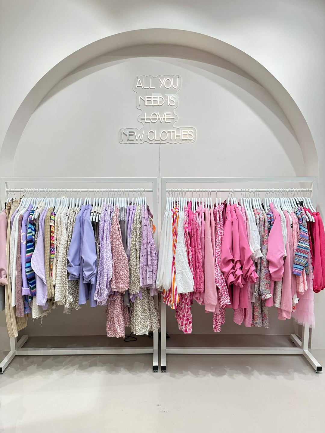 Bunter Kleiderständer mit pastellfarbener Damenmode in Rosa, Flieder und Blumenmustern in Lisa's Boutique unter dem Leuchtspruch "All you need is love – and new clothes"