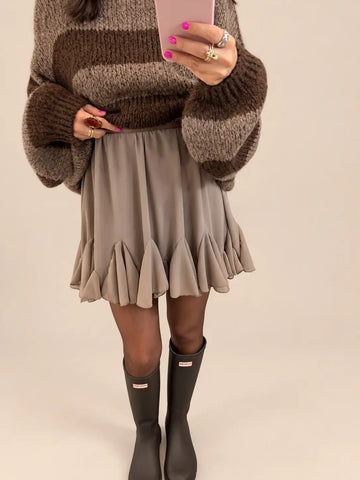 Falda corta color topo para mujer con volante en el bajo, con jersey de rayas y botas sobre fondo de estudio beige
