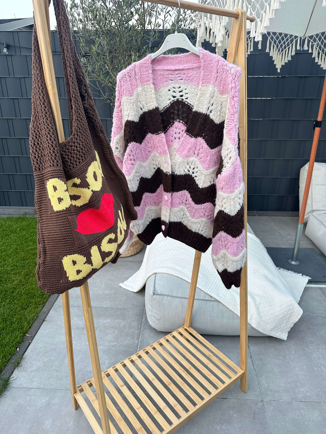 Strickjacke in Braun-Rosa-Weiß mit Zickzack-Muster, hängend an einer Kleiderstange neben einer braunen Häkel-Tasche mit gelber Aufschrift „BISO“ und rotem Herz.