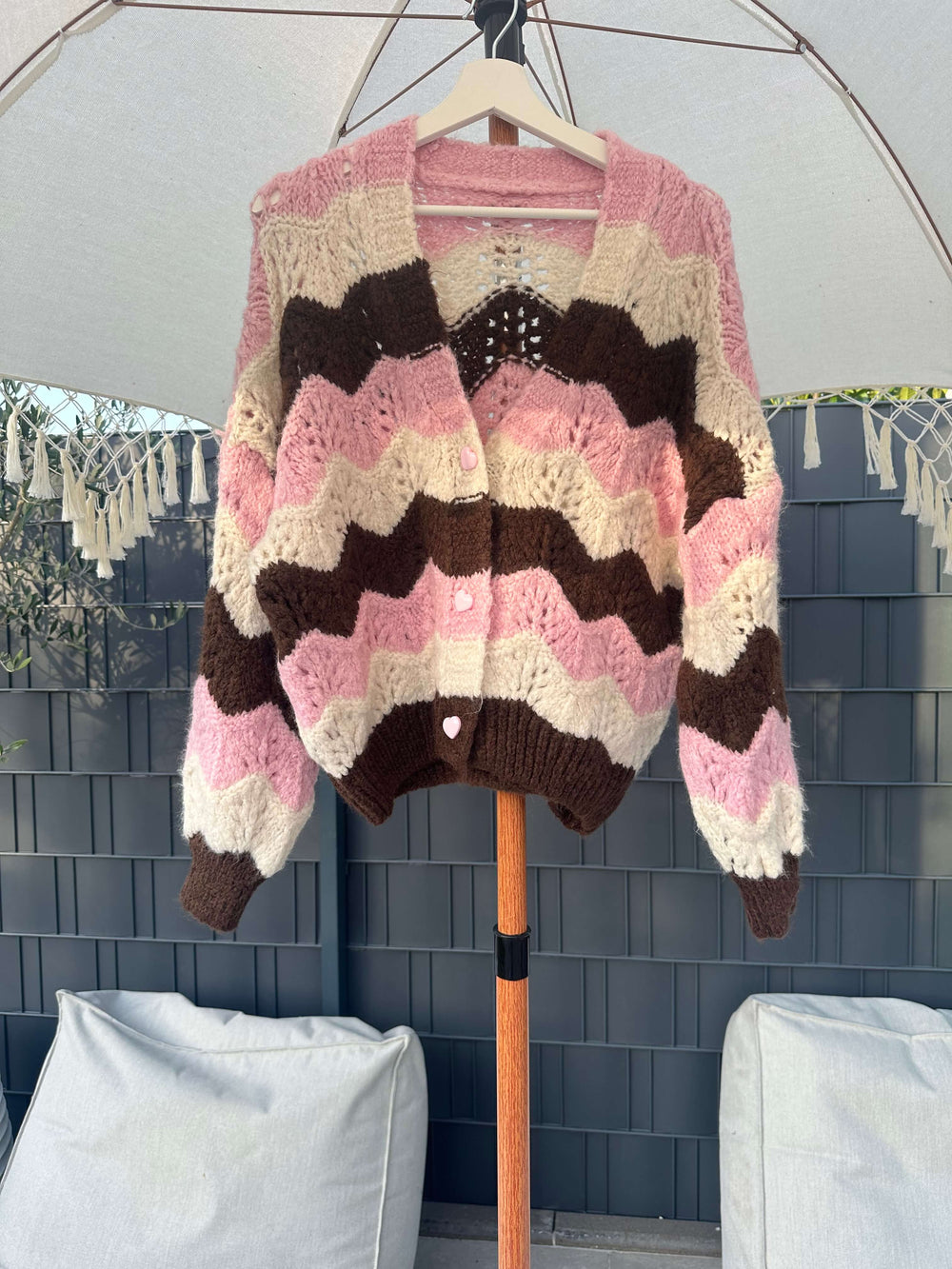 Kuschelige Strickjacke mit braun-rosa-weißen Zickzack-Streifen, hängend auf einem Bügel unter einem Sonnenschirm im Gartenbereich.