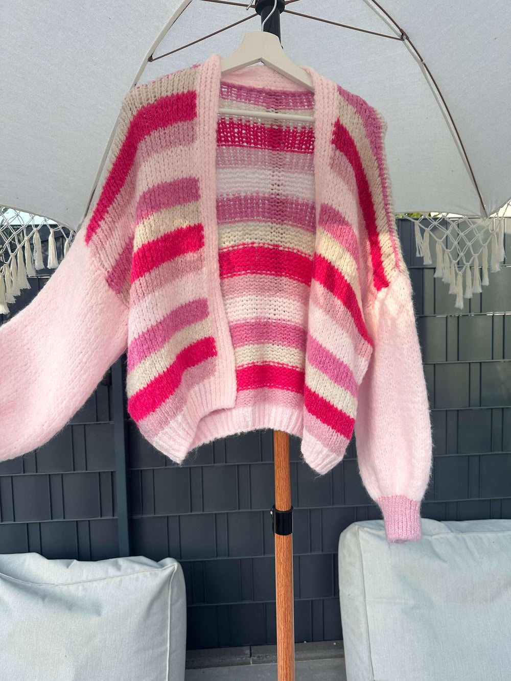 Strickjacke mit breiten Streifen in Rosa-, Pink- und Gelbtönen, hängend auf einem Kleiderbügel unter einem Sonnenschirm im Outdoor-Bereich.