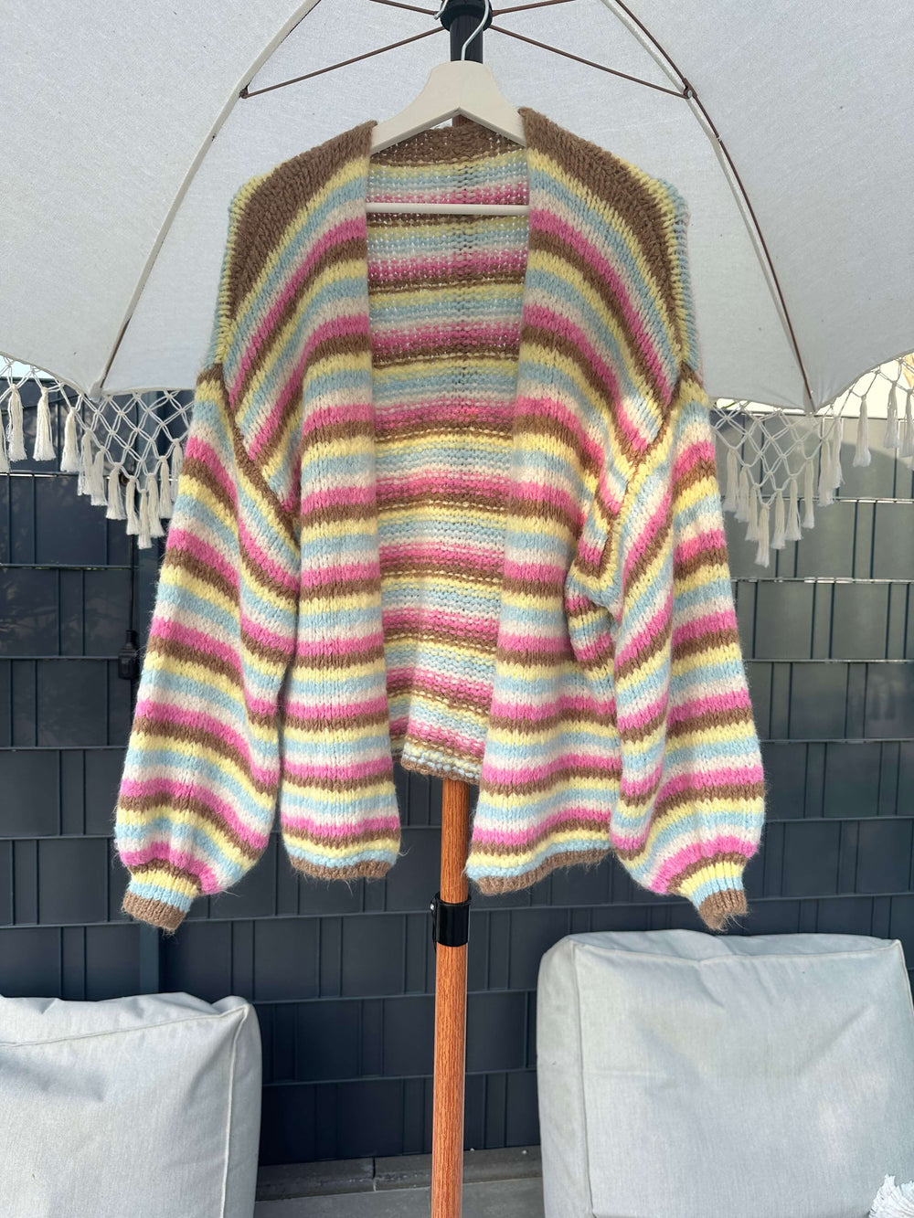 Strickjacke mit mehrfarbigen Streifen in Braun-, Gelb-, Pink- und Blautönen, hängend auf einem Kleiderbügel unter einem Sonnenschirm im Outdoor-Bereich.