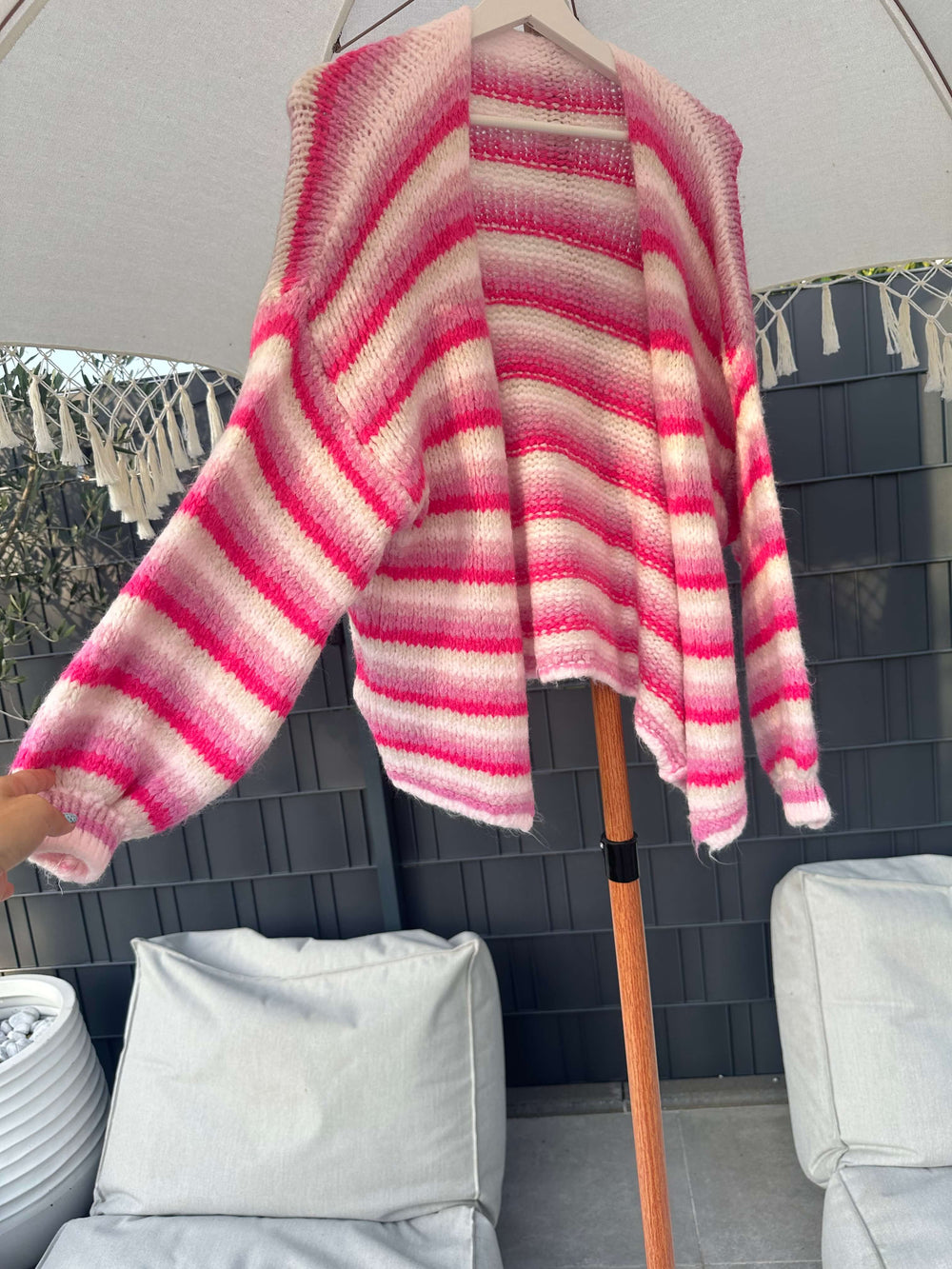 Strickjacke in Rosa-Weiß mit breiten Querstreifen, hängend auf einem Kleiderbügel unter einem Sonnenschirm im Outdoor-Bereich.